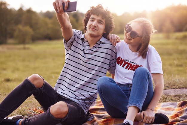 Urocza Para Baw Się Razem, Usiądź Na Kratce Na świeżym Powietrzu, Ciesz Się Słoneczną, Ciepłą Pogodą, Pozuj Do Robienia Selfie Z Nowoczesnego Smartfona. Kędzierzawy Męski Nastolatek Robi Fotografii Z Komórkowym. Letni Odpoczynek