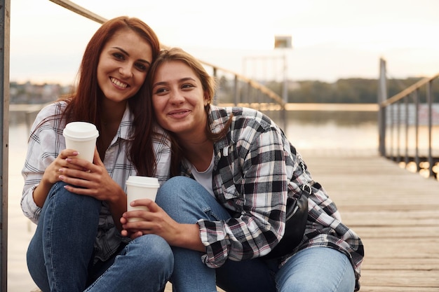 Urocza para lesbijek siedzi razem nad jeziorem z kubkami napoju w dłoniach i obejmuje się nawzajem