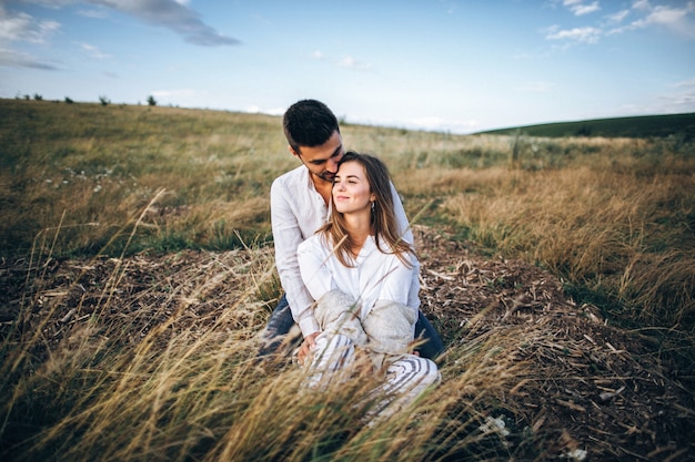 Urocza para przytulająca się, całująca i uśmiechająca się na tle nieba siedząca na trawie