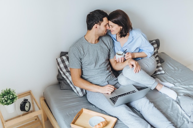 Urocza para romantyczna zamierzająca się pocałować patrzeć na siebie siedząc na łóżku używać laptopa do oglądania filmów pić kawę cieszyć się wspólnotą i relaksem w sypialni Koncepcja związku