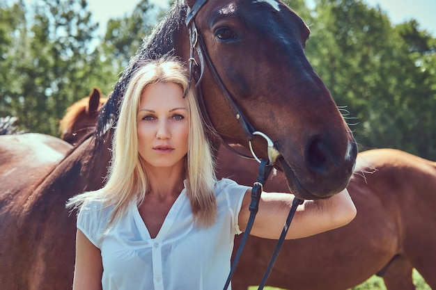 Urocza piękna blondynka w białej bluzce i dżinsach przytula konia do zielonego pola.
