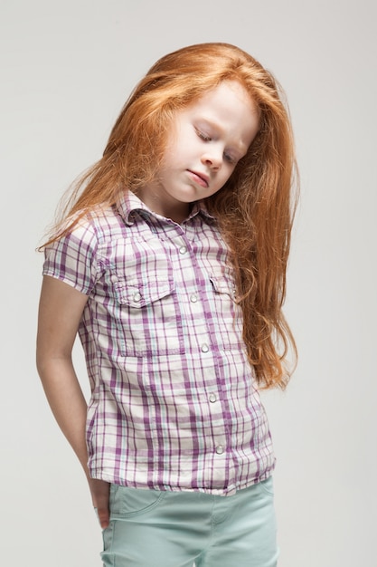 Urocza rudowłosa dziewczynka w kraciastej koszuli, jasnoniebieskich spodniach i białych butach