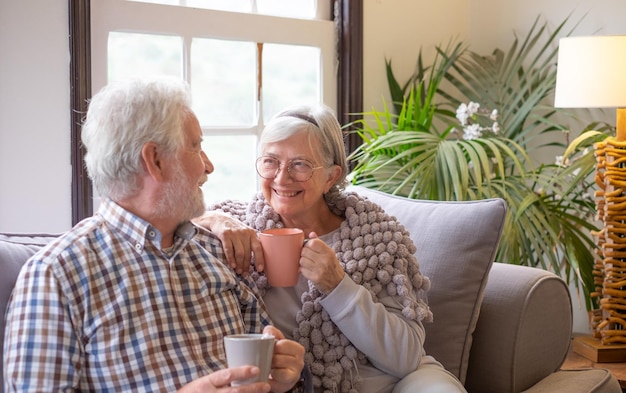 Urocza starsza para relaksuje się na kanapie w salonie, popijając filiżankę herbaty lub kawy, ciesząc się emeryturą i wolnym czasem