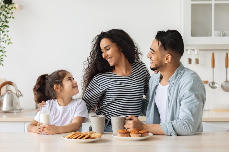 Urocza szczęśliwa arabska rodzina je śniadanie podczas wspólnego weekendu w domu, wesoły młody ojciec, mama i kędzierzawa dziewczynka siedzą w kuchni, pijąc kawę i mleko z domowymi ciasteczkami