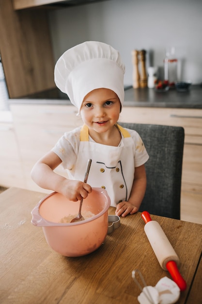 Urocza, szczęśliwa dziewczyna w szefowym kapeluszu i fartuchu gotuje w kuchni.
