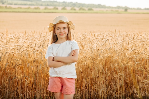 Urocza szczęśliwa dziewczynka w kapeluszu w polu pszenicy patrząc w kamerę ze skrzyżowanymi rękami ...