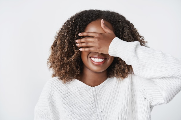 Urocza, szczęśliwa i zachwycona urocza afrykańsko-amerykańska koleżanka blisko wzroku dłonią i spodziewająca się prezentu w ciepłym swetrze na białej ścianie