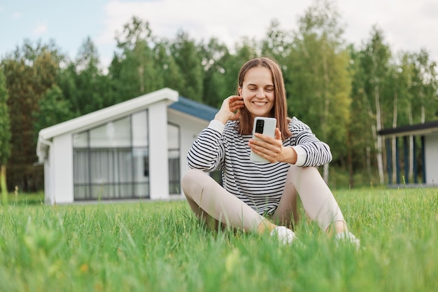 Urocza urocza kobieta w parku z domem w tle, przeglądająca internet na smartfonie, mająca rozmowę wideo, robiąca selfie rozmawiająca z przyjacielem w internecie