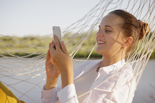 Urocza uśmiechnięta kaukaska kobieta w białej koszuli siedzi w hamaku na brzegu rzeki i używa telefonu komórkowego, wpisując wiadomości lub sprawdzając sieci społecznościowe