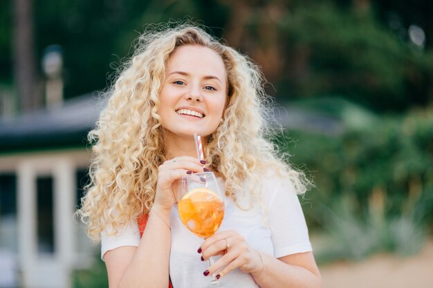 Urocza wesoła blondynka o kręconej kobiecie z pozytywnym wyrazem, pijąca zimny letni koktajl, spacerująca na świeżym powietrzu, chętnie otrzymująca komplement od chłopaka, dobrze odpoczywająca. Ludzie, rozrywka, styl życia