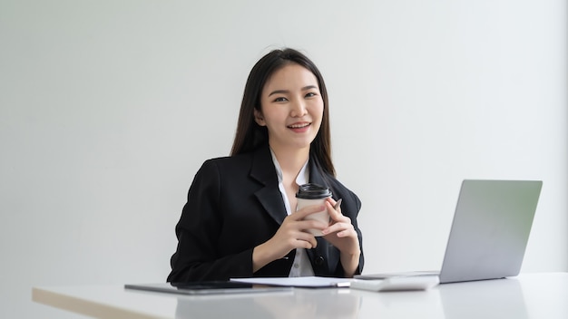 Urocze azjatyckie businesswoman siedzi w biurze gospodarstwa dokumentu laptopa filiżanka kawy. Patrząc na aparat.
