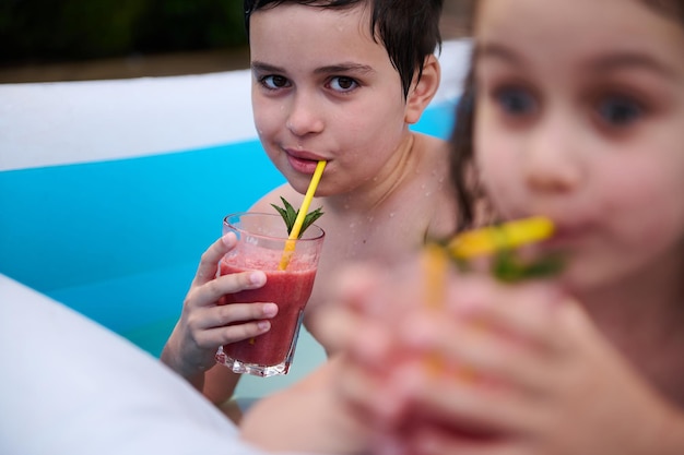 Urocze dzieci odpoczywają w nadmuchiwanym basenie z wodą i piją świeżo wyciśnięty sok owocowy ze słomki do picia