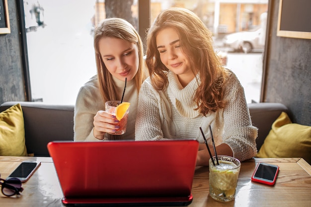 Urocze Młode Kobiety Siedzą Przy Stole I Patrzą Na Laptopa. Oni Oglądają Film. Modele Mają Okulary Przeciwsłoneczne, Napoje I Telefony Przy Stole. Jeden Z Nich Trzyma W Rękach Szklankę Koktajlu.