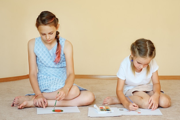 Urocze siostry siedzące na podłodze z papierami i rysujące akwarelami w domu