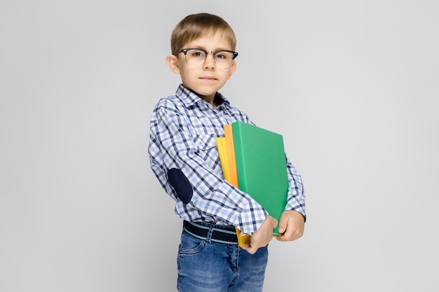 Zdjęcie uroczy chłopiec w koszuli vkletchatoy i jasnych dżinsach stoi na szaro. chłopiec trzyma w rękach wielobarwny folder z dokumentami
