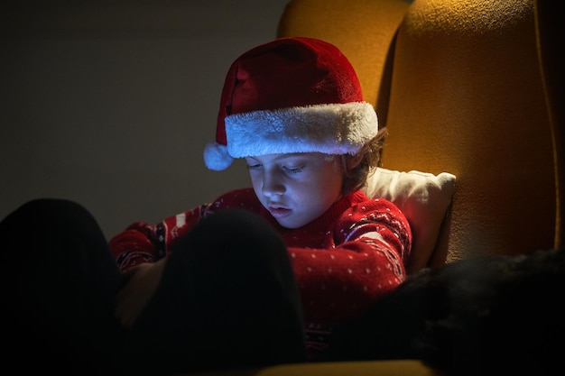 Uroczy, ciekawski chłopiec w czapce Mikołaja i swetrze korzystający z tabletu, siedząc wieczorem w żółtym fotelu w mieszkaniu