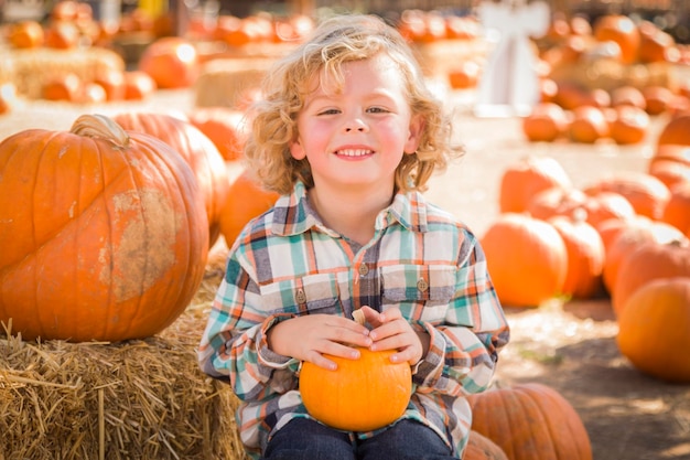 Uroczy mały chłopiec siedzi i trzyma dynię na rustykalnym ranczu na Pumpkin Patch