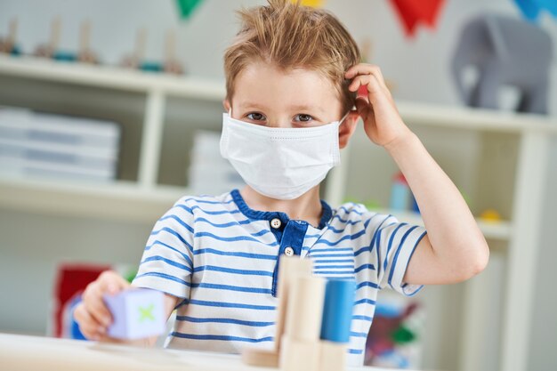 Uroczy mały chłopiec w przedszkolu z założoną maską z powodu pandemii koronawirusa