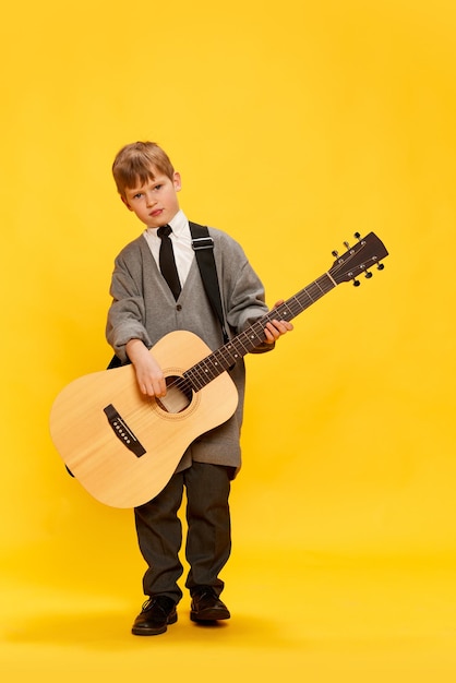 Uroczy mały chłopiec, zabawny muzyk, ubrany w ogromne ubrania oversize, grający na gitarze, dobrze się bawiący na białym tle