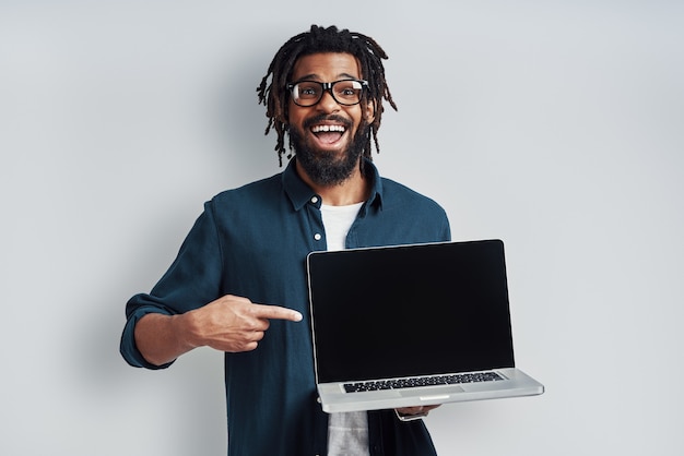 Uroczy młody mężczyzna w okularach wskazujący miejsce na kopię na laptopie, stojący przy szarej ścianie