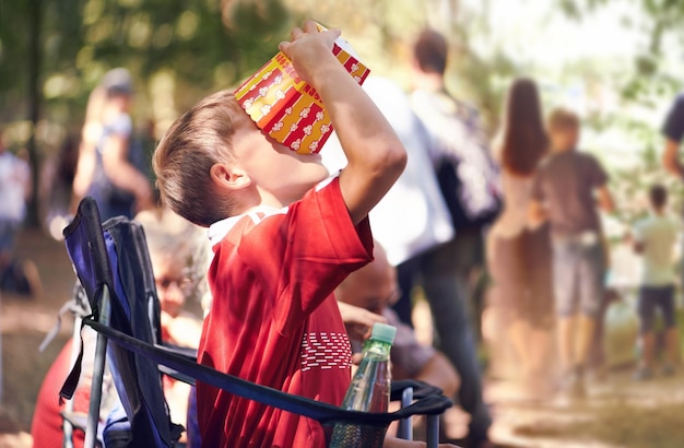 Uroczystość festiwalowa i chłopiec jedzący popcorn na świeżym powietrzu w lecie na uroczystości dla świętowania lub rozrywki Jedzenie dla dzieci lub przekąska i małe dziecko jedzące z pudełka w przyrodzie las lub las sam na krześle