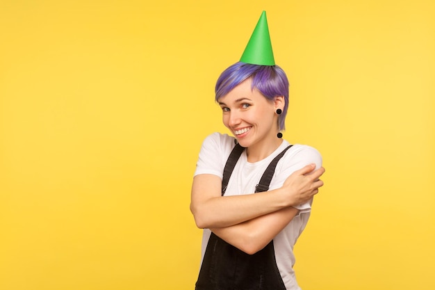 Urodziny portret samolubnej hipster dziewczyny z fioletowymi krótkimi włosami na sobie zabawny stożek strony i kombinezon, obejmując się i uśmiechniętą samoocenę koncepcja na białym tle na żółtym tle strzał studio