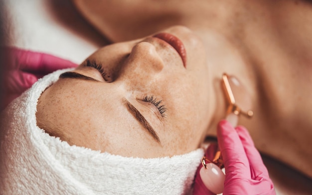 Zdjęcie urządzenie toczące się do detoksykacji doskonałej skóry reklamy produktów do pielęgnacji skóry spa wellness zbliżenie portret kobiety otrzymującej masaż twarzy za pomocą kamiennego wałka gua sha w salonie spa
