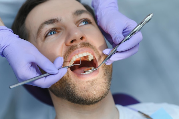 Usługi periodontologiczne Zbliżenie strzał uśmiechający się mężczyzna leczenie w klinice stomatologicznej Lekarz dentysta w rękawiczkach za pomocą sterylnych narzędzi dentystycznych do badania zębów pacjenta płci męskiej