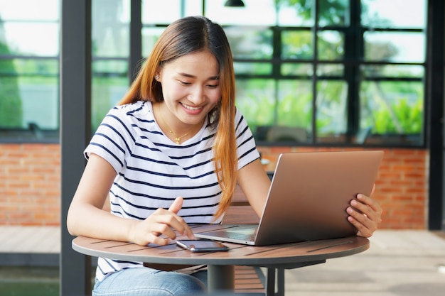 Uśmiecha się azjatykciej dziewczyny używa wiszącą ozdobę i laptop przy kawiarnią