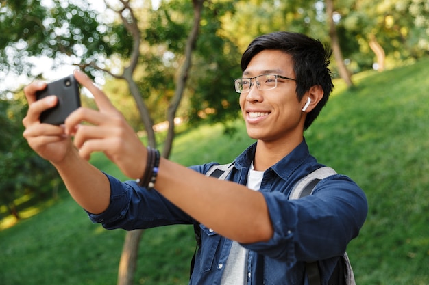 Uśmiechający się azjatycki student płci męskiej w okularach robi selfie na smartfonie będąc w parku