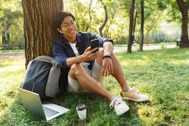 Uśmiechający Się Azjatycki Student Płci Męskiej W Okularach Za Pomocą Swojego Smartfona Siedząc W Pobliżu Drzewa W Parku