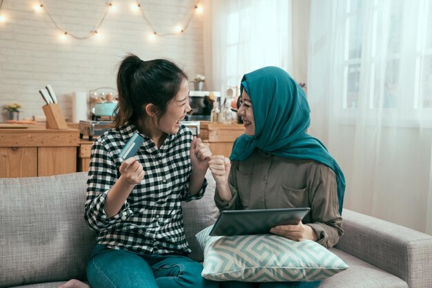 Uśmiechający się azjatyckich przyjaciół za pomocą cyfrowego tabletu i karty kredytowej w domu. współlokatorka azjatyckie kobiety islamskie i koreańskie kupujące w sklepie internetowym letniej wyprzedaży. podekscytowane panie roześmiane i pusty koszyk na zakupy