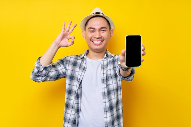 Uśmiechający się fajny przystojny Azjatycki młody mężczyzna 20s na sobie przypadkowe ubrania kapelusz pokazujący pusty ekran telefonu komórkowego i ok, w porządku gest na białym tle na żółtym tle portret studyjny Ludzie koncepcja stylu życia