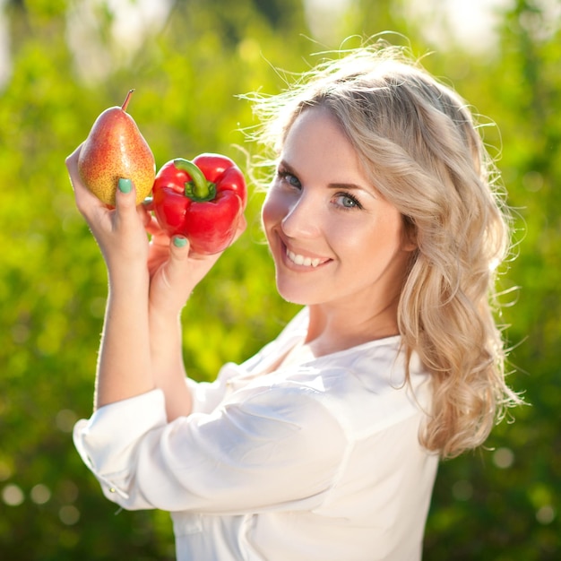 Uśmiechający się piękna blond kobieta trzyma owoce i warzywa na zielonym tle przyrody.