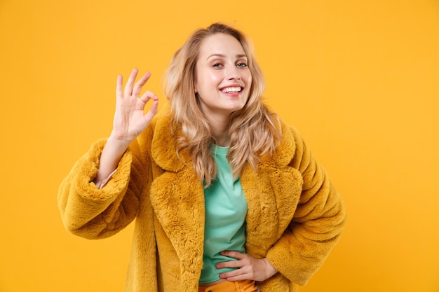 Uśmiechający się piękna młoda blondynka kobieta dziewczyna w żółtym futrze pozowanie na białym tle na pomarańczowym tle portret studio. Koncepcja życia szczere emocje ludzi. Makieta miejsca na kopię. Pokazuje OK gest.