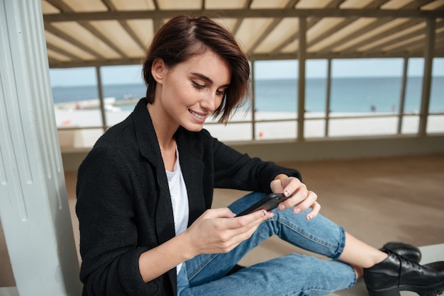 Uśmiechający Się Uroczy Młoda Kobieta Siedzi I Korzystania Z Telefonu Komórkowego W Altanie Na Plaży