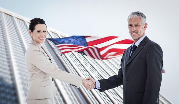 Uśmiechnięci ludzie biznesu uściskają dłonie, patrząc w kamerę na amerykańską flagę i wieżowiec