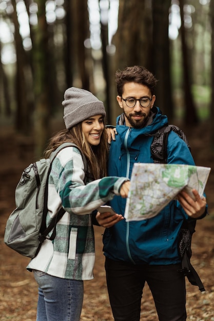 Uśmiechnięci młodzi Europejczycy turyści w kurtkach z plecakiem spacerują po jesiennym lesie patrzą na mapę