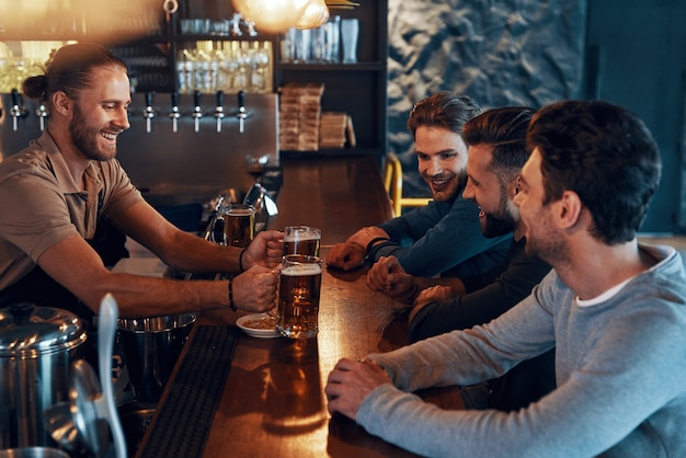 Uśmiechnięci młodzi mężczyźni w codziennych ubraniach, pijący piwo i nawiązujący więź, siedząc w pubie