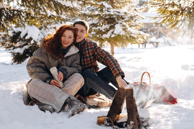 Uśmiechnięci nowożeńcy siedzą przy małym ogniu w środku śnieżnego, słonecznego parku. Młody mężczyzna i