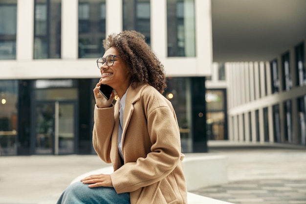 Uśmiechnięta afro amerykańska bizneswoman rozmawia przez telefon siedząc na tle nowoczesnego budynku
