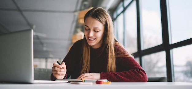 Uśmiechnięta atrakcyjna hipster dziewczyna odrabiająca pracę domową, sprawdzająca informacje za pomocą nowoczesnego laptopa