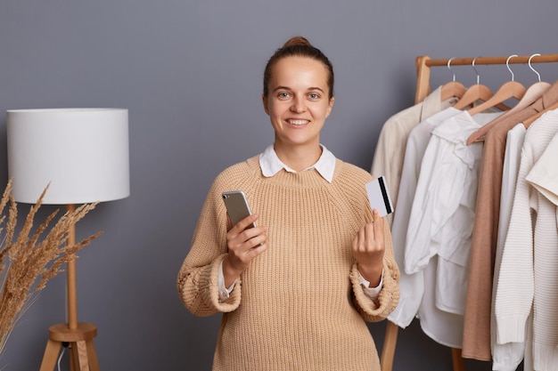 Uśmiechnięta atrakcyjna kobieta z fryzurą kok, ubrana w beżowy sweter stojący ze smartfonem i kartą kredytową płacąca za stroje online, pozowanie na szarej ścianie z ubraniami na wieszakach na półce