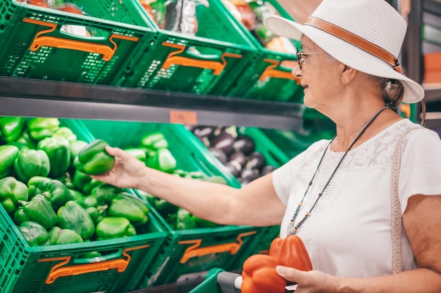 Uśmiechnięta atrakcyjna starsza klientka w kapeluszu wybierająca warzywa w supermarkecie trzymająca pieprz