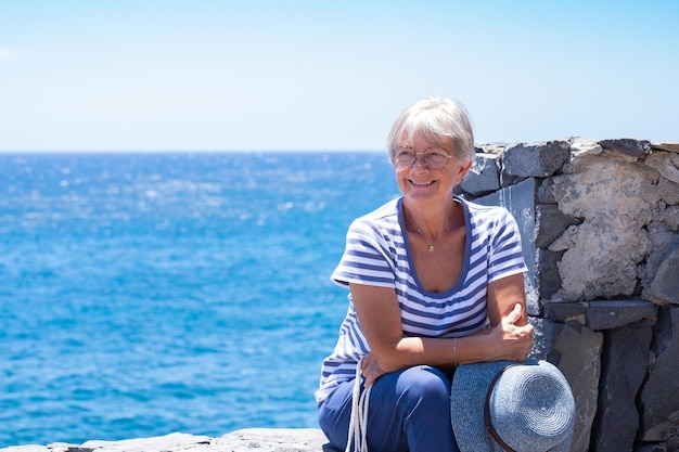 Uśmiechnięta atrakcyjna starsza kobieta ubrana na niebiesko, siedząca blisko morza w letnie wakacje, ciesząc się słonecznym dniem