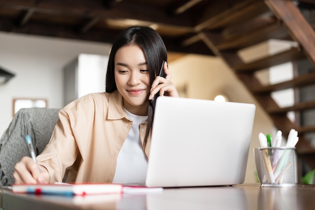 Uśmiechnięta Azjatka odbiera telefon rozmawiając przez komórkę i zapisując notatki pracując na laptopie od...