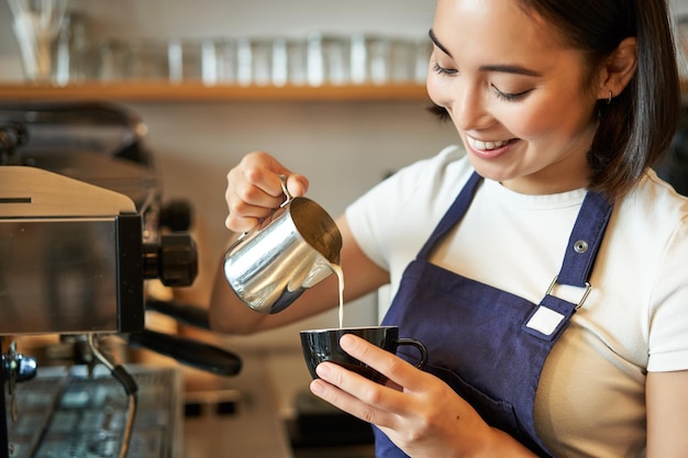 Uśmiechnięta azjatycka barista dziewczyna robi kawie wlewając mleko na parze do cappuccino, robiąc latte art w filiżance