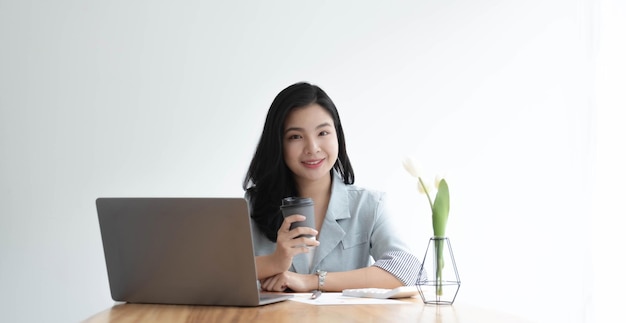 Uśmiechnięta azjatycka bizneswoman trzymająca kubek kawy i laptopa w biurze Patrząc na kamerę