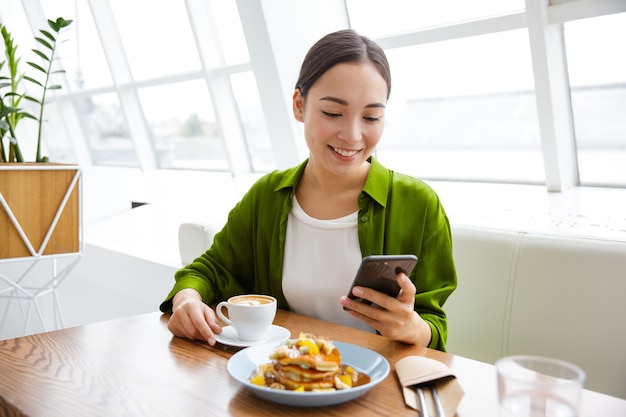 Uśmiechnięta Azjatycka Kobieta Je Naleśniki Na śniadanie W Kawiarni W Pomieszczeniu, Trzymając Telefon Komórkowy