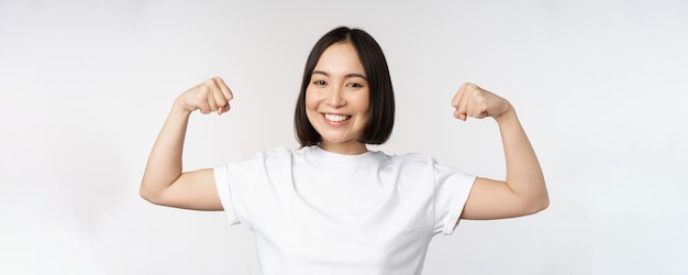 Uśmiechnięta azjatycka kobieta pokazująca napinające mięśnie bicepsa silny gest ramion stojący w białej koszulce na białym tle